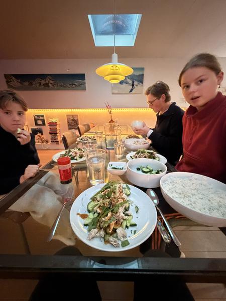 This photo captures three people enjoying a casual dinner around a glass dining table, illuminated by a distinctive yellow pendant lamp